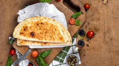 calzone with salami, ham, cheese, tomato, basil, mushrooms, cream sauce on a wooden background, Italian pastries, traditional Italian cuisine, olive oil with spices, food photo, flatlay