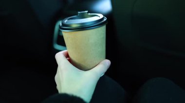 Bir kadın eli, arabanın arka koltuğunda siyah plastik kapaklı kahverengi bir eko-bardak tutuyor. Arabanın içi odak noktasında, yolcu koltuğunun arkası görünür durumda.. 