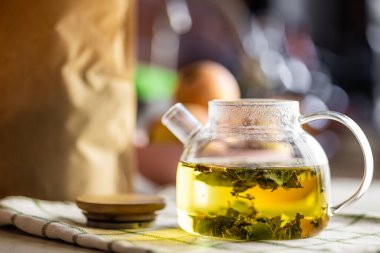 Yeşil yapraklı bitkisel çay içeren temiz bir cam çaydanlık, kontrol edilmiş bir bez üzerinde dinleniyor. Işık kehribar sıvısı güneş ışığına hafifçe yansıyarak şeffaf, yuvarlak şekli vurgular.