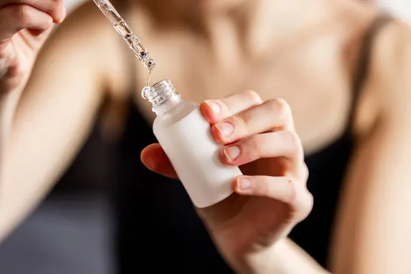 Elinde buzlu cam şişeyle yüz serumu tutan bir kadın. Şişe silindirik ve metal bir damlacıkla kaplanmış. Pipetten bir damla serum akar. Kincare ya da kozmetik uygulama kavramı