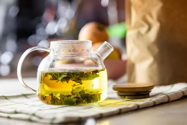 Yeşil yapraklı bitkisel çay içeren temiz bir cam çaydanlık, kontrol edilmiş bir bez üzerinde dinleniyor. Işık kehribar sıvısı güneş ışığına hafifçe yansıyarak şeffaf, yuvarlak şekli vurgular.