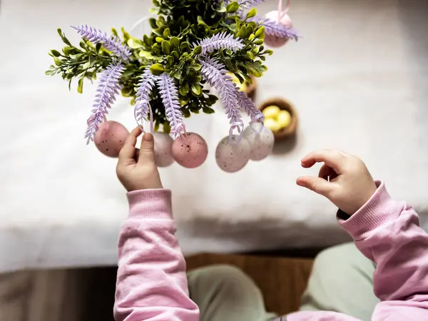Çocuk, boyalı Paskalya yumurtalarıyla bir dalı süslüyor, bir çocukla Paskalya için hazırlanıyor, dekoratif bir yuvada pembe ve sarı bıldırcın ve tavuk yumurtası boyuyor.