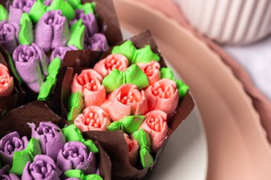 Beyaz bir tabakta lale şeklinde şekerli meyveli ve kremalı kekler, Anneler Günü için ev yapımı pastalar, hafif kompozisyon, arka planda çay, lale ve pembe ipek kurdeleli kekler.