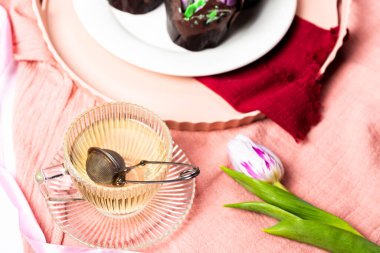 Hafif arka planda çay fincanı olan cam bir fincan çay, beyaz tabakta lale şeklinde şeker ve kremalı muffinlerin yanında, Anneler Günü için ev yapımı pastalar, beyaz laleli vazolar.