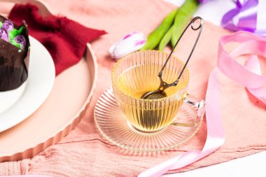 Hafif arka planda çay fincanı olan cam bir fincan çay, beyaz tabakta lale şeklinde şeker ve kremalı muffinlerin yanında, Anneler Günü için ev yapımı pastalar, beyaz laleli vazolar.