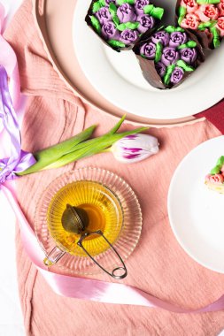 Beyaz bir tabakta lale şeklinde şekerli meyveli ve kremalı kekler, Anneler Günü için ev yapımı pastalar, hafif kompozisyon, arka planda çay, lale ve pembe ipek kurdeleli kekler.