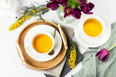 Beyaz porselen fincanlar ve çaydanlıkta yeşil çay, cam vazoda laleler, mimoza çiçekleri, açık arkaplan. Çay partisi..