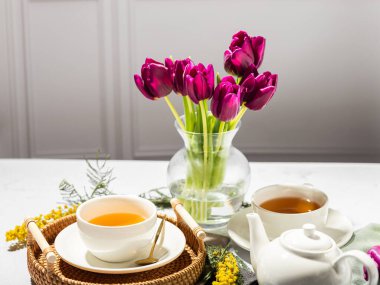 Beyaz porselen fincanlar ve çaydanlıkta yeşil çay, cam vazoda laleler, mimoza çiçekleri, açık arkaplan. Çay partisi..