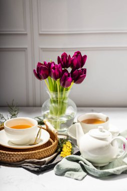 Beyaz porselen fincanlar ve çaydanlıkta yeşil çay, cam vazoda laleler, mimoza çiçekleri, açık arkaplan. Çay partisi..