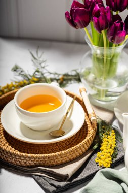 Beyaz porselen fincanlar ve çaydanlıkta yeşil çay, cam vazoda laleler, mimoza çiçekleri, açık arkaplan. Çay partisi..