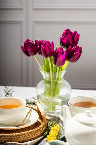 Beyaz porselen fincanlar ve çaydanlıkta yeşil çay, cam vazoda laleler, mimoza çiçekleri, açık arkaplan. Çay partisi..