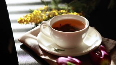 Siyah arka planda, güneş ışığında yeşil çay içilen beyaz bardak. Beyaz porselen bardak ve çay tabağı. Yakında sarı çiçekler var. Yüksek kaliteli video