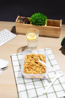 Ofis ortamı, yemek molası. İş yerinde atıştırmalık bir şeyler. Çizgili bir kumaşa konulmuş havuçlu ve tavuklu kızarmış pirinç konteynırı. Limonlu bir bardak su. Yakın klavye