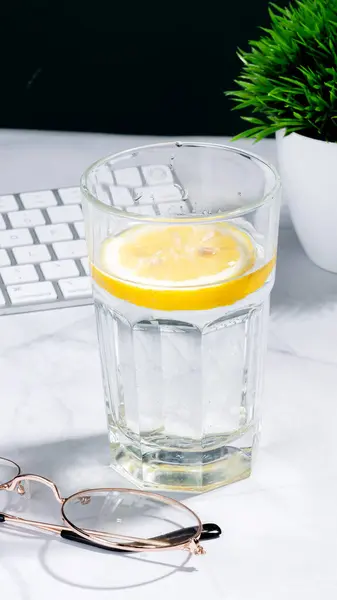 Su dolu berrak bir bardak ve ışık masasında limon dilimi. Yakınlarda, beyaz bir bilgisayar tuş tahtası. Ofis ortamı, dinlendirici bir mola ya da iş sırasında susuz kalma girişiminde bulunulduğunu gösteriyor.
