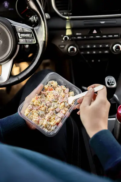 Arabadaki beslenme çantasından salata yiyen adam. Arabada atıştırmalık bir şeyler var. Şoför koltuğundaki adam öğle yemeğinde salata yiyor..
