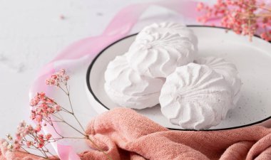 Pembe kurdele ve çiçeklerin yanında beyaz bir tabakta beyaz şekerleme zephyr. Elma püresi ve yumurta beyazından yapılmış beyaz zefyr. Düşük kalorili tatlılar. Vegan şekerlemesi..