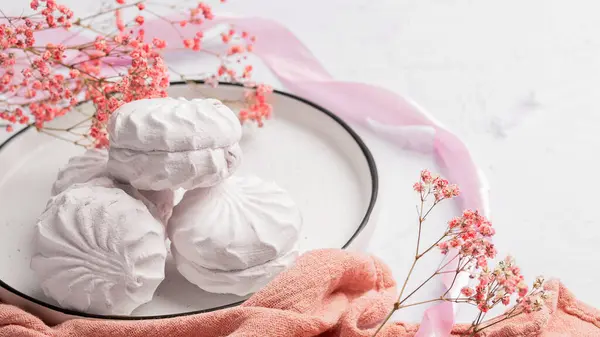 Pembe kurdele ve çiçeklerin yanında beyaz bir tabakta beyaz şekerleme zephyr. Elma püresi ve yumurta beyazından yapılmış beyaz zefyr. Düşük kalorili tatlılar. Vegan şekerlemesi..