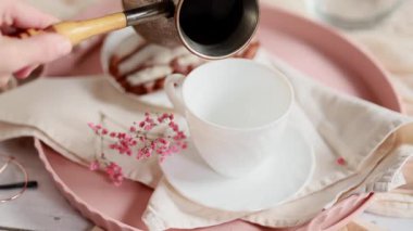 Bir kadın kahve fincanından çay tabağıyla beyaz bir fincana kahve dolduruyor. Taze demlenmiş sade kahve buharlaşıyor. Hafif arkaplan. Pembe çiçekler. Kremalı pasta yakınlarda..