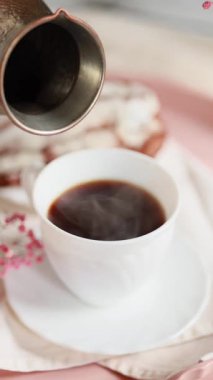 Bir kadın kahve fincanından çay tabağıyla beyaz bir fincana kahve dolduruyor. Taze demlenmiş sade kahve buharlaşıyor. Hafif arkaplan. Pembe çiçekler. Kremalı pasta yakınlarda..