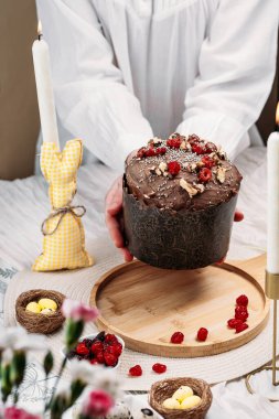 Beyaz gömlekli kadın Paskalya pastası, çikolata kaplı paskalya ekmeği kuru kiraz ve cevizle süslenmiş arka planda uzun mumlar, çiçekler, Paskalya tavşanı. Paskalya konsepti.