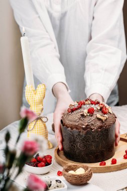 Beyaz gömlekli kadın Paskalya pastası, çikolata kaplı paskalya ekmeği kuru kiraz ve cevizle süslenmiş arka planda uzun mumlar, çiçekler, Paskalya tavşanı. Paskalya konsepti.