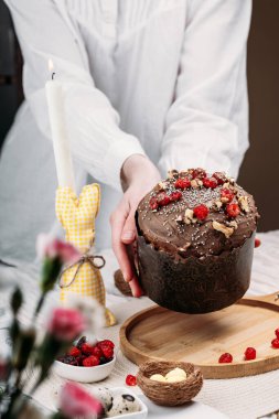 Beyaz gömlekli kadın Paskalya pastası, çikolata kaplı paskalya ekmeği kuru kiraz ve cevizle süslenmiş arka planda uzun mumlar, çiçekler, Paskalya tavşanı. Paskalya konsepti.