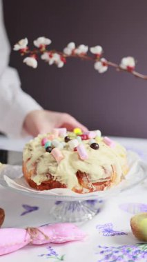 Beyaz gömlekli kadın tereyağı kremalı ve şekerli büyük bir Cinnabon kesiyor. Paskalya ekmeği. Çiçekler ve Paskalya dekoru yakında.