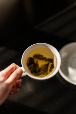 Bir el, çay yapraklı yeşil çay fincanını tutarken, beyaz bir çay tabağının üzerine yerleştirilmiş. Koyu arkaplan kontrastı güçlendirir, çayların tazeliğini ve organikliğini vurgular