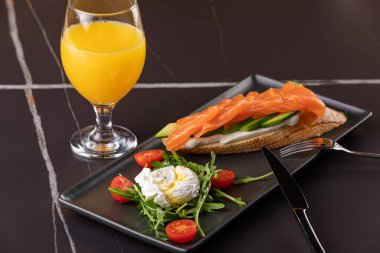 brxchetta with avocado and salted salmon. On a plate with poached egg. In the frame of a glass with orange juice and a plate with bruscheta. black background. Close-up.
