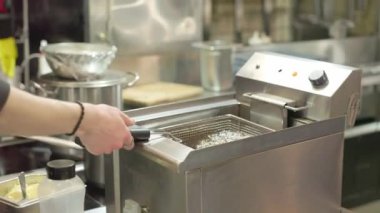 Pişirme yağının üstündeki patates kızartmasıyla sepeti sallıyor. Konteynırı patates kızartmasıyla birlikte kızgın yağın içine yerleştiriyorum. Patates kızartması sıcak yağda çabucak pişirilir. Fast Food. Kızartma makinesi