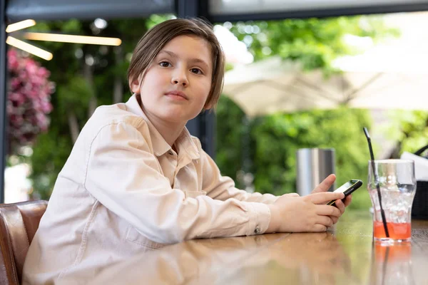 stock image Young cheerful teen playing on mobile phone sitting behind a table in a park or outdoor cafe. child is sitting in a garden, on a sunny summer day. boy playing on a cell phone outdoor.