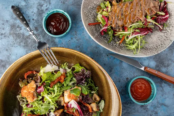 İki salatanın üst görüntüsü. Dilimlenmiş biftekli yeşil yapraklı biftek salata ve taze salata tavuk göğsü, roka ve gri taş arka planda mavi salata.