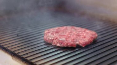 Üç hamburger ızgarada pişiyor, alevler köftelerin kenarlarını yalıyor. Hamburgerler kahverengileşiyor ve cızırdıyor, ağız sulandıran bir koku yaratıyor.