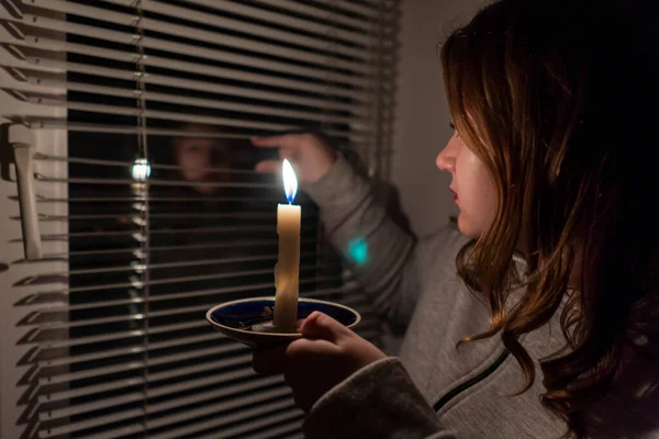 Kesinti. Enerji krizi. Altyapı tahribatı. Elektrik kesintisi konsepti. Pencerenin yanındaki karanlık bir odada elinde yanan bir mum tutan bir kız.