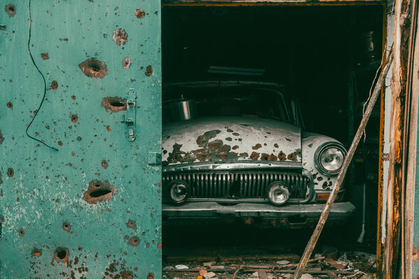 Сломанная гражданская винтажная машина в гараже. На кузове автомобиля дырки от пуль и осколков. Война на Украине. Русское вторжение на Украину. Военные преступления