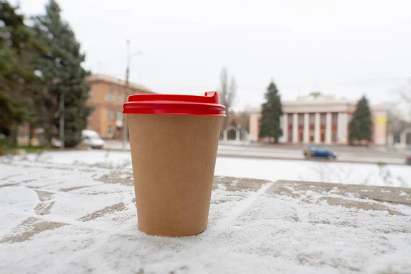 Winter. Cold weather. The ground is covered with a small layer of snow. There is a cup of coffee on the ground against the background of a city street (close-up)