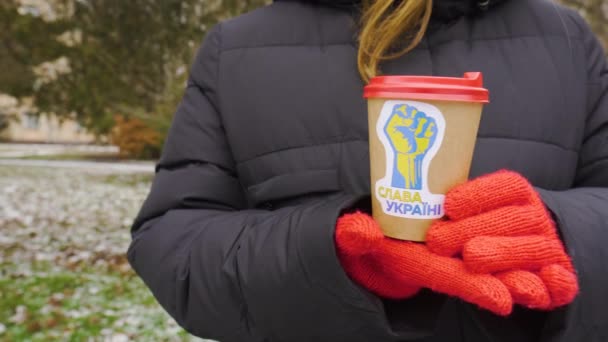 寒冷的天气城市公园 一个女孩手里拿着一杯咖啡 手里拿着爱国的贴纸 固定相机 贴纸上题词的翻译 荣耀归于乌克兰 — 图库视频影像