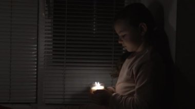 A girl in a dark room by the window holds a candle in a glass in her hands (fixed camera). Power outage concept. Blackout. Energy crisis. Destruction of infrastructure