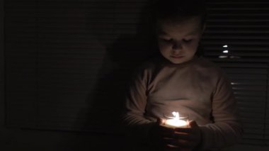 A girl in a dark room by the window holds a candle in a glass in her hands (close-up, the camera pans from left to right). Power outage concept. Blackout. Energy crisis. Destruction of infrastructure