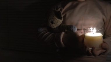 A girl in a dark room by the window holds a candle and a teddy bear in her hands (close-up, the camera pans from left to right). Power outage concept. Blackout. Energy crisis