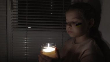 A girl with a painted flag of Ukraine on her cheek in a dark room holds a candle in her hands (fixed camera). Power outage concept. Blackout. Energy crisis. Destruction of infrastructure