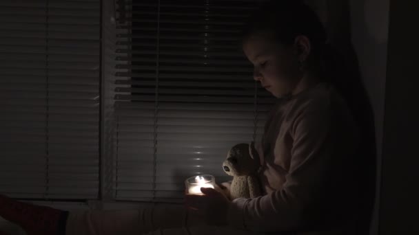 一个女孩在窗边一间黑暗的房间里 手里拿着一支蜡烛和一只泰迪熊 相机从左向右移动 停电的概念 停电了能源危机 基础设施被毁 — 图库视频影像