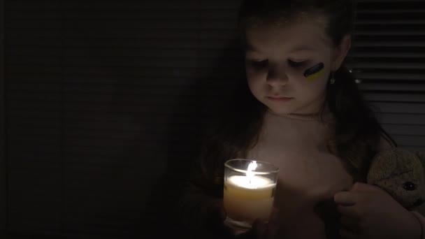 在一间黑暗的房间里 一个脸上挂着乌克兰国旗的女孩手里拿着一支蜡烛和一只泰迪熊 相机从左到右都有 停电的概念 停电了 — 图库视频影像