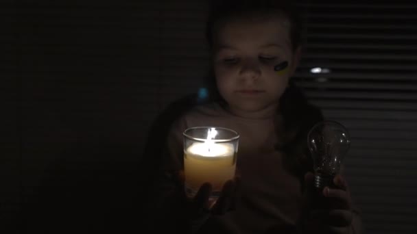 在一间黑暗的房间里 一个脸上挂着乌克兰国旗的女孩手里拿着一支蜡烛和一个电灯泡 相机从左到右都有 停电的概念 停电了 — 图库视频影像