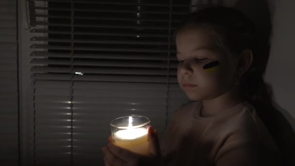 在黑暗的房间里 一个脸上挂着乌克兰国旗的女孩手里拿着蜡烛 固定相机 停电的概念 停电了能源危机 基础设施被毁 — 图库视频影像