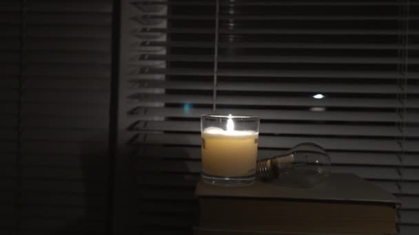 灯泡躺在窗边的一间黑暗的空房间里 旁边点着一支蜡烛 特写镜头 摄像机从左到右转动 停电了能源危机 停电的概念 — 图库视频影像