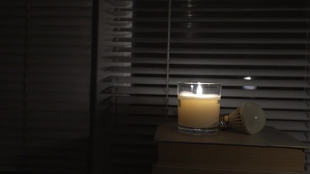 Led灯泡躺在窗边的一间黑暗的空房间里 旁边点着一支蜡烛 特写镜头 摄像机从左到右转动 停电了能源危机 停电的概念 — 图库视频影像