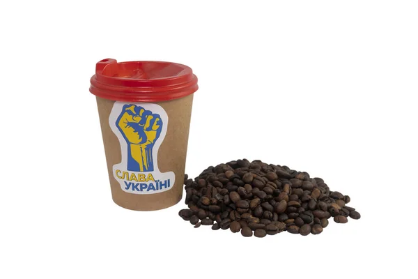 A paper cup of coffee with a patriotic sticker and a handful of roasted coffee beans isolated on a white background (close-up). Translation of the text on the sticker - 