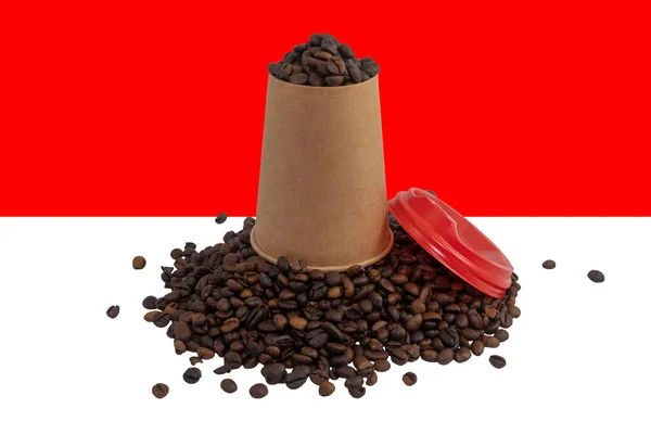 An overturned disposable paper cup with a lid filled with roasted coffee beans against the background of the Indonesian flag (close-up). The largest exporter of coffee in the world. Indonesian coffee