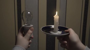 A man in a dark room holds a holder with a light bulb in one hand and a burning candle in the other hand (close-up, fixed camera). Blackout. Power outage concept